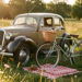 Voiture ancienne garée dans une prairie au coucher du soleil, accompagnée d’un vélo rétro, d’un panier de pique-nique et d’une couverture à carreaux.