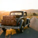 Voiture vintage chargée de bagages en cuir roulant sur une route de campagne au coucher du soleil.