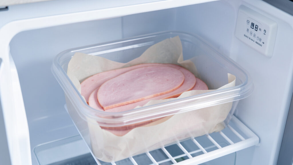Tranches de jambon blanc séparées par du papier cuisson dans une boîte hermétique placée au congélateur.