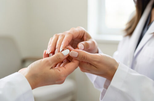 Gros plan sur la main d’une femme aux ongles pâles et blanchâtres examinée avec douceur par une médecin dans un cabinet médical.