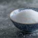 Bol de sucre semoule blanc posé à côté d’une cuillère doseuse sur un plan de travail.
